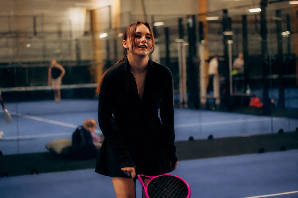 Uma jovem sorri em uma quadra de padel com sua raquete fúcsia na mão.