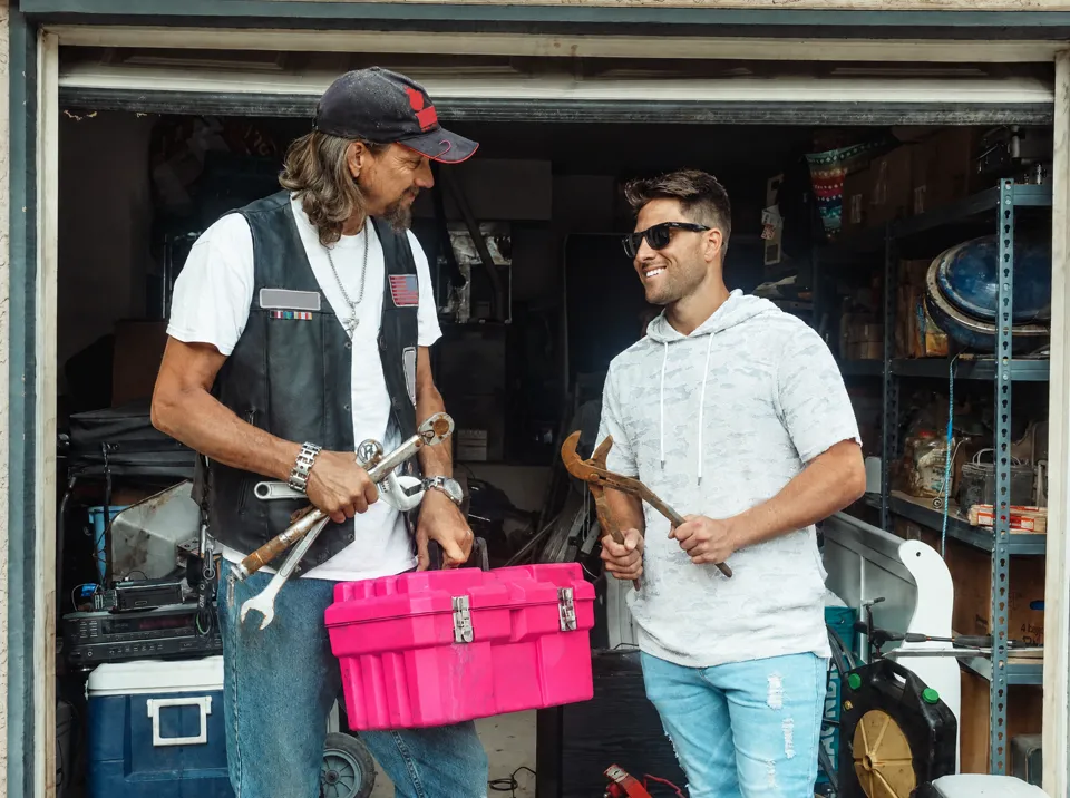 Un mecánico con chaleco de cuero sostiene llaves y una caja de herramientas fucsia en la puerta de su taller.
