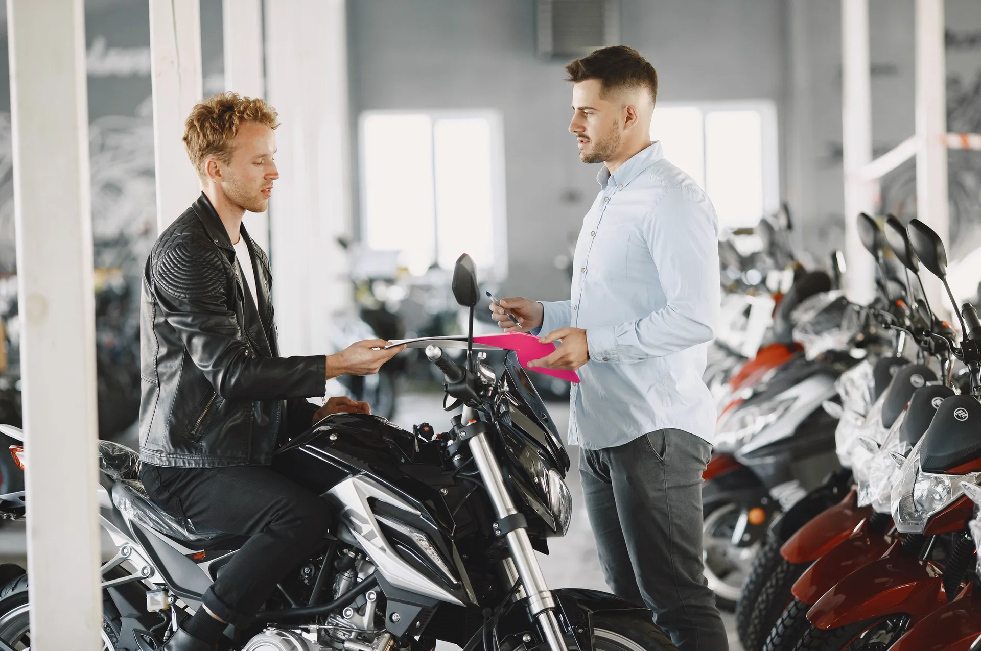 Un vendedor con una carpeta fucsia atiende a un cliente en un concesionario multimarca, con varias motos de fondo.